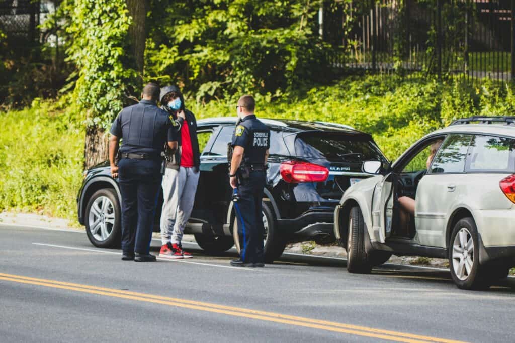 Police officers talking to a person next to a parked black SUV on the side of the road, with another open vehicle beside them.